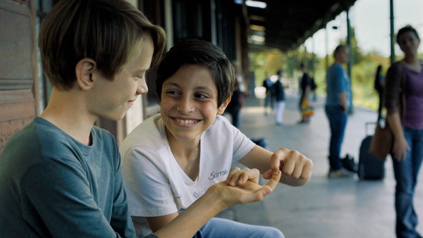 Zwei Jungen sitzen am Bahnsteig und grinsen einander an.