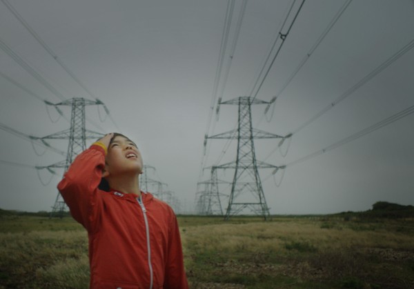 Ein kleiner Junge steht auf einem Feld und blickt, die Hand am Kopf, hinauf zu den Stromleitungen.