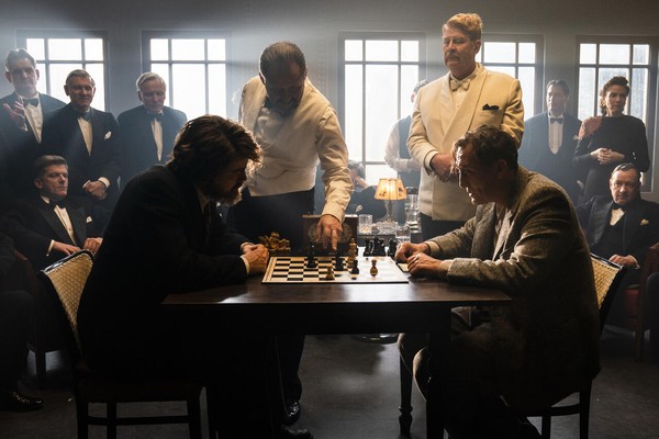 Zwei Männer sitzen an einem Tisch und spielen Schach vor einem elegant gekleideten Publikum.