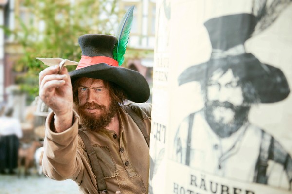 Der Räuber Hotzenplotz schaut hinter einer Litfaßsäule hervor, auf der sein Fahndungsplakat klebt, und wirft einen Papierflieger.