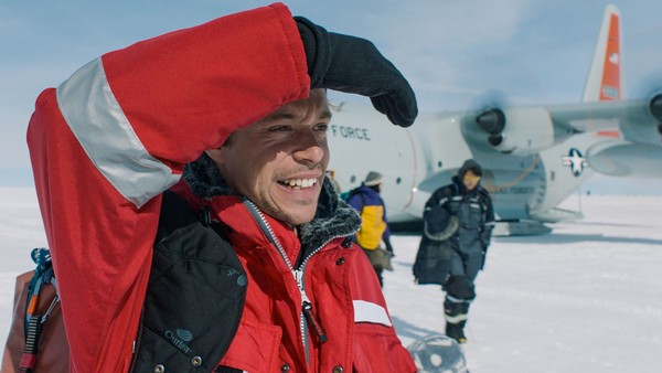 Ein Mann mit Winterjacke und Rucksack steht im Schnee und blickt in die Ferne. Hinter ihm stehen ein Flugzeug und zwei Menschen.