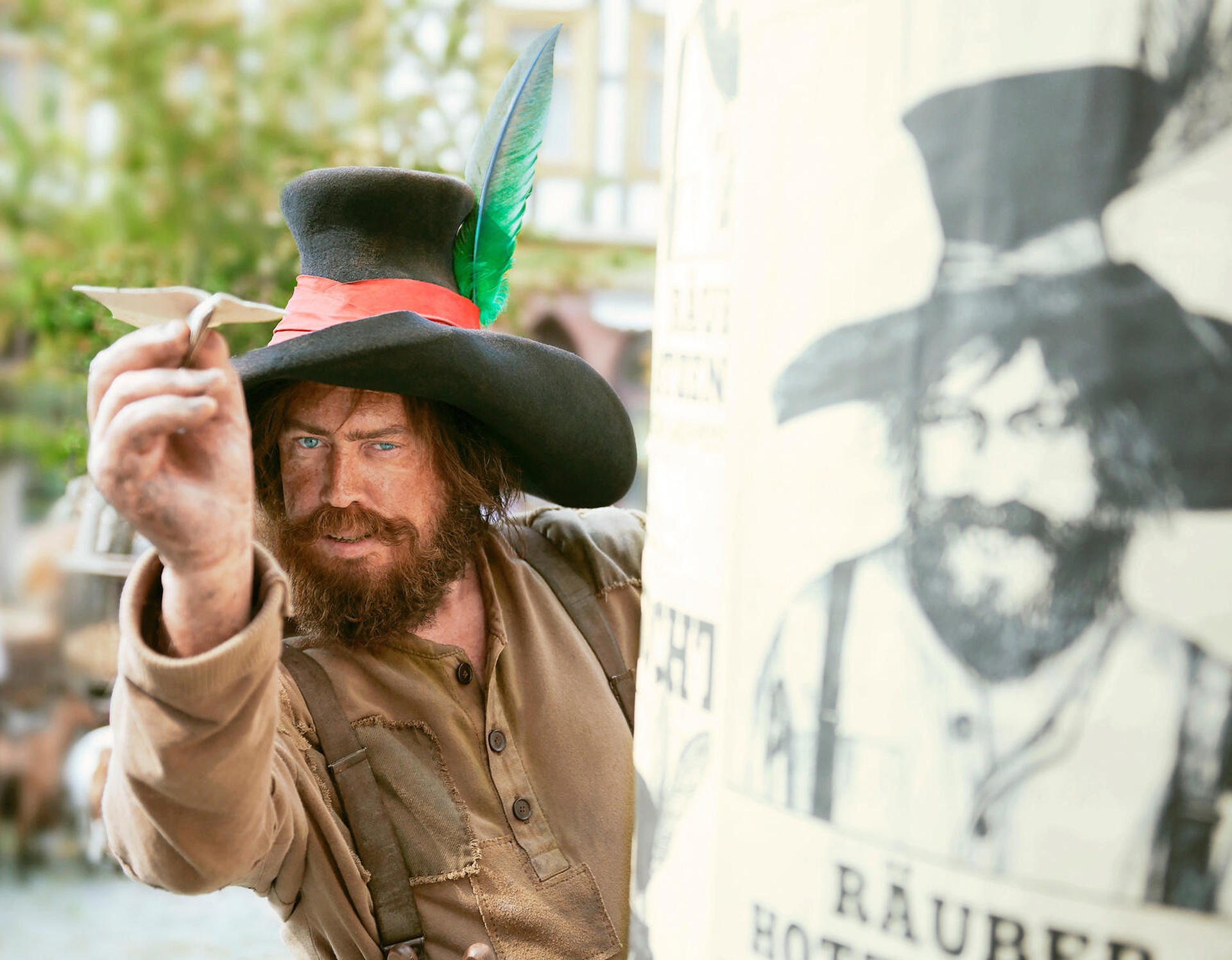 Der Räuber Hotzenplotz schaut an einer Litfaßsäule vorbei, auf der ein Fahndungsplakat von ihm hängt, und wirft einen Papierflieger.