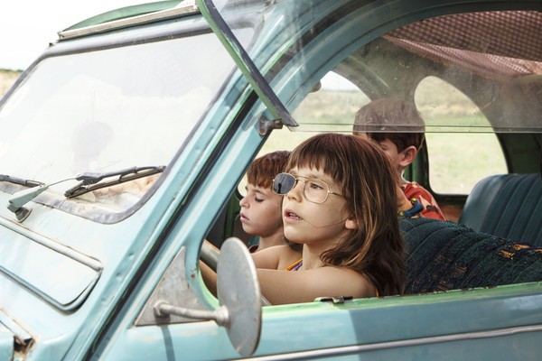 Drei Kinder sitzen in einem Autowrack, das Mädchen am Steuer trägt eine Sonnenbrille, die aber nur noch ein Glas hat