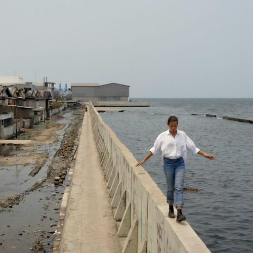 Eine junge Frau balanciert auf einer Mauer, links von ihr das Meer, rechts von ihr eine Häusersiedlung, die überschwemmt wurde.