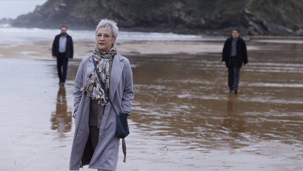 Eine elegant gekleidete Frau spaziert den Strand entlang, im Hintergrund ihre Leibwächter.