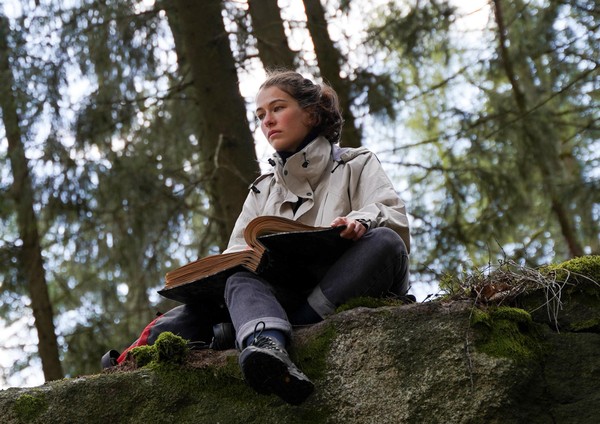 Eine junge Frau sitzt im Wald auf einer Steinkante mit einem Buch auf dem Schoß.