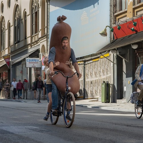 Ein Mädchen fährt mit ihrem als Wurst verkleideten Vater zusammen auf dem Fahrrad durch die Stadt.