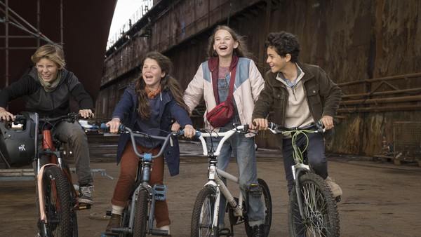Vier Kinder sitzen lachend auf ihren Fahrrädern inmitten alter Schiffswracks.