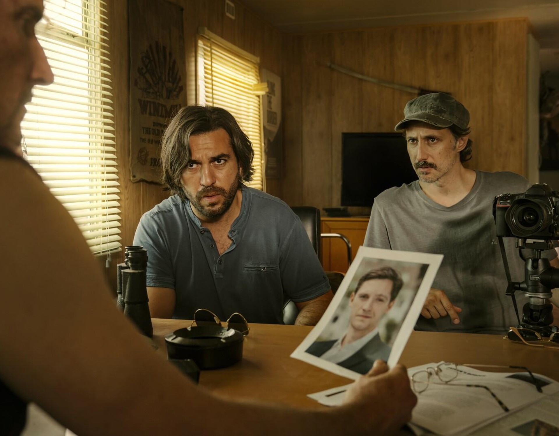Zwei Journalisten sitzen in einem Büro und präsentieren einem dritten ein Bild des Mannes, den sie suchen.