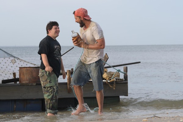 Ein junger Mann mit Down-Syndrom und ein bärtiger Mann mit einer Flasche Schnaps in der Hand stehen im seichten Wasser vor einem Floß und lachen